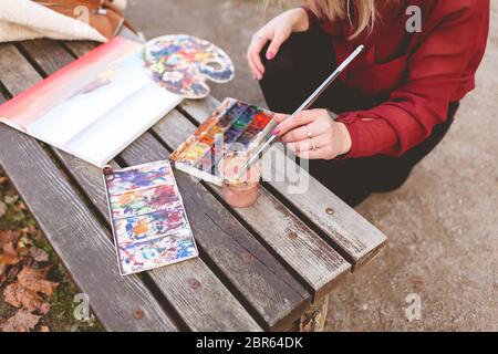 Frau zeichnet ein Bild auf einer Parkbank. Blonde Künstlerin engagiert sich in ihrem Lieblings-Hobby des Zeichnens. Zeichenzubehör Stockfoto