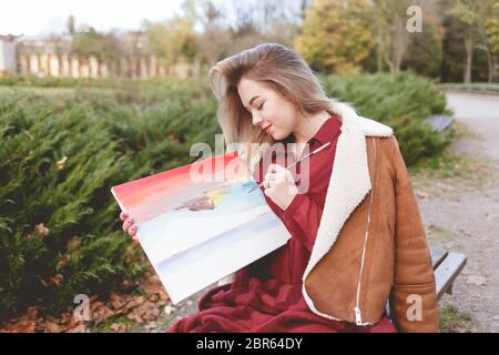 Das junge Mädchen zeichnet ein Bild auf einer Parkbank. Blonde Künstlerin engagiert sich in ihrem Lieblings-Hobby des Zeichnens Stockfoto