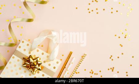 Goldene Party Dekorationen auf beigefarbenem Hintergrund Draufsicht. Flache Lay Einkaufstasche mit Geschenk, Trinkhalmen, goldenem Band und Konfetti. Weihnachten, Geburt Stockfoto