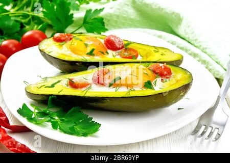 Rührei mit Tomaten in zwei Hälften von Avocado in einem Teller, grün Handtuch und Gabel auf dem Hintergrund der weißen Holzbrett Stockfoto