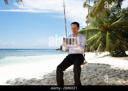 Mann sitzt auf Schaukel mit digitalen Tablet am Sandstrand in der Nähe der idyllischen Meer Stockfoto