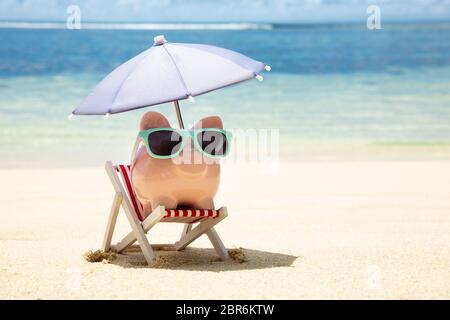 Rosa Sparschwein mit Sonnenbrille und Sonnenschirm auf Liegestuhl auf Sand am Strand Stockfoto