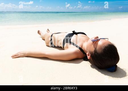 Junge Frau im schwarzen Bikini, Sonnenbrille, entspannend am Sandstrand in der Nähe des Meeres Stockfoto