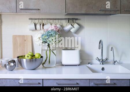 Küche in beigefarbenen Tönen. Becher und Milchkännchen hängen an Haken. Waschbecken mit Wasserhähnen. Stahlschale mit grünen Äpfeln. Bouquet von bunten Hortensien in einem Glas Stockfoto