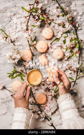 Flach-legen von Frauen Hände halten süße Macaron Cookies und frischen Kaffee in Tasse über weißen Frühlingsblüten Blumen und weißem Marmor Hintergrund, Draufsicht Stockfoto