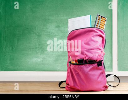 Pinker Rucksack mit Buch und verschiedenem Briefpapier auf dem Holztisch mit Kreidetafel Hintergrund. Back to School Konzept Stockfoto