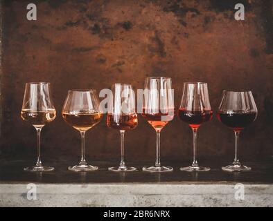 Verschiedene Schattierungen von Roséwein in stieligen Gläsern in Linie von hell bis dunkel auf Beton Tisch, rostigen braunen Hintergrund hinter, kopieren Raum platziert. Stockfoto