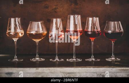 Verschiedene Schattierungen von Roséwein in gestielten Gläsern in Linie von hell bis dunkel auf Beton Tisch, rostbraun Hintergrund hinter gelegt. Weinbar, wi Stockfoto