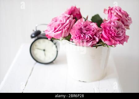 Ein Strauß rosa Rosen in einem Zinn weißen Eimer auf einem alten Hocker mit Wecker und einer Serviette mit Stickerei neben dem Bett auf einem beigen Hintergrund Stockfoto