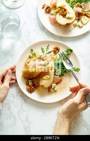 Weibliche Hände halten einen Teller Birnensalat mit Walnüssen, Prosciutto, Rucola und Blauschimmelkäse. Abendessen Vorspeise auf Marmortisch Stockfoto