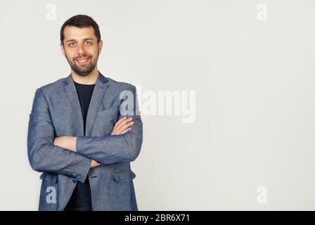 Porträt eines jungen männlichen Unternehmers lächelnd breit mit einem selbstbewussten Ausdruck, Geschäftsmann lächelnd mit gekreuzten Armen. Mann, der auf die Kamera schaut, isoliert über einer grauen Wand mit Kopierraum. Stockfoto