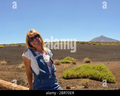 Eine Frau, die auf einem Kofferraum sitzt, Sonnenbrille trägt, weißes Hemd, blaue kurze Latzhose, hat blonde lange Haare, im Hintergrund ein prächtiges vulkanisches Land Stockfoto