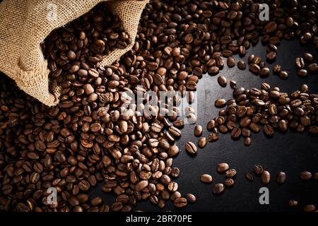 Geröstete Kaffeebohnen mit kleinen Sackleinen Tasche in die Ecke Auslaufen auf einen dunklen Schiefer Oberfläche mit Highlight gesehen hohen Winkel Stockfoto