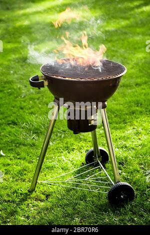 Flaming heißes Feuer in einen tragbaren Grill im Freien auf einem üppigen, grünen Rasen mit einem leeren Grill in der Nähe zu Seitenansicht Stockfoto