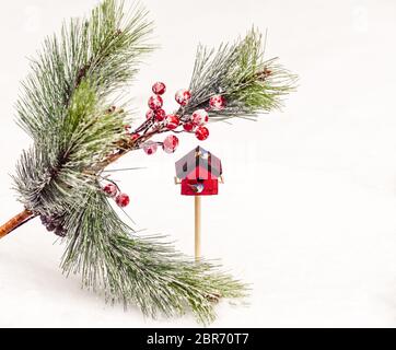 Frisch gefallener Schnee im Hintergrund mit gefälschten Kiefernästen, Beeren und zwei Vögeln auf einem winzigen roten Vogelhaus thront Stockfoto