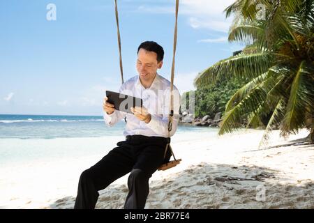 Mann sitzt auf Schaukel mit digitalen Tablet am Sandstrand in der Nähe der idyllischen Meer Stockfoto