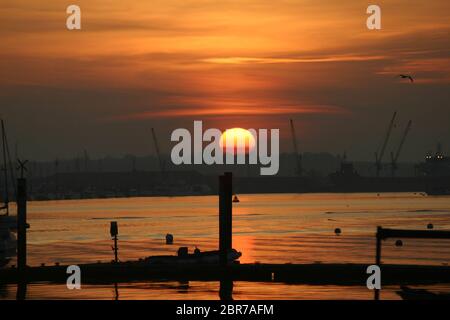 Leuchtender orangefarbener Sonnenuntergang über dem Fluss Crouch in Burnham auf Crouch in Essex. Im Vordergrund stehen Stege und Boote mit Kräne und Frachtschiffen Stockfoto