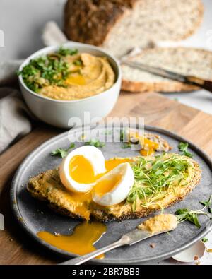 Ein weich gekochtes Ei, in die Hälfte auf einer dünnen Scheibe Brot mit Hummus und Grüns auf einem silbernen Teller, der auf einem Holzbrett sitzt, mit einer Schüssel bedeckt geschnitten Stockfoto