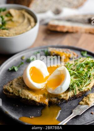 Ein weiches gekochtes Ei in der Nähe auf einer dünnen Scheibe Brot, die mit Hummus und Grüns mit einer Schüssel Hummus und dem Laib Brotscheiben bedeckt ist, halbieren Stockfoto