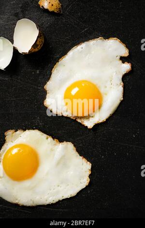 2 gebratene Wachteleier auf schwarzer Kochfläche mit rissigen Eierschalen zur Seite. Stockfoto