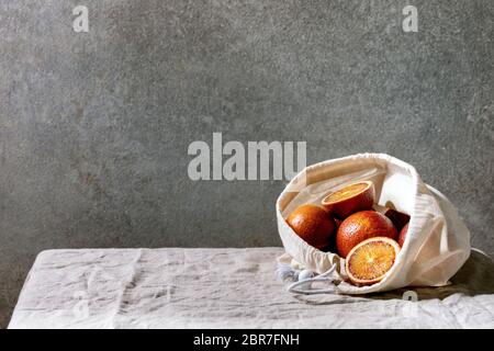 Blut sizilianische Orangen, reif und saftig, in Baumwolle umweltfreundliche Tasche, ganz und in Scheiben geschnitten, auf grauem Leinen Tischtuch, Betonwand als Hintergrund. Stockfoto