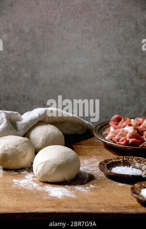 Teig für italienische Pizza napolitana Kochen. Drei Kugeln aus frischem, selbstgemachtem Weizenteig, Prosciutto und Zutaten in Keramikplatten oben auf Holz-KI Stockfoto