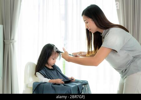 Asiatische Mutter Haare schneiden ihre Tochter im Wohnzimmer zu Hause während zu Hause bleiben sicher von Covid-19 Coronavirus während der Sperrung. Selbstquarantäne an Stockfoto