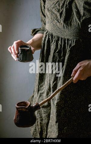 Frau in dunklem Kleid halten in Händen Handwerk handgemachte Keramik-Kaffeekanne cezve mit langen Holzgriff und Kanne Milch für die Herstellung von türkischen Kaffee. Stockfoto