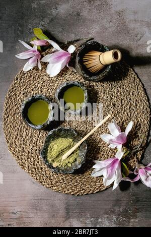 Japanischer Matcha-Grüntee in handgemackten Keramiktassen mit Matcha-Pulver, Bambusbesen und rosa Magnolienblüten auf Strohserviette über dunklem Textur-Backgroun Stockfoto