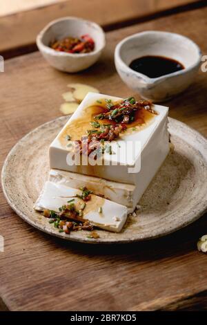 Seidentofu japanischer Sojakäse Vollstück mit Chili Ingwer, Chive und Sojasoße, die auf Keramikplatte über Holztisch aufliegen. Stockfoto