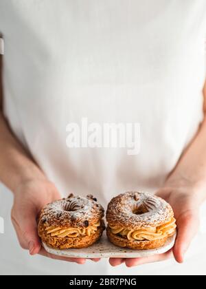 Frau in weißem T-Shirt hält Teller mit zwei Choux Paris Brest Gebäck mit Haselnuss Stockfoto