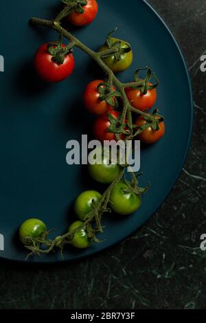 Kirschtomaten auf der Rebe, einige reif und einige unreif, auf einem blaugrünen Teller, auf einem grünen Marmorgrund. Stockfoto
