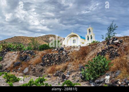 Typische Landschaft der Insel Santorini an einem Sommer bewölkten Tag. Auf dem Hügel im Dorf Akrotiri befindet sich eine traditionelle weißblaue orthodoxe Kirche Stockfoto