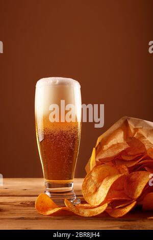 Schließen Sie ein Glas Lagerbier mit weißen Schaum und Blasen und Papier Tüte Kartoffelchips auf hölzernen Tisch über dunkelbraunen Hintergrund mit Kopie Raum Stockfoto