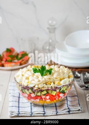 Große Glasschüssel mit Krabbbensalat, Mais, Gurke und Reis am Tisch. Mehrschichtige Krabbenstäbchen und Maissalat. Traditiolnal Mayonnaise Salat für russische holi Stockfoto