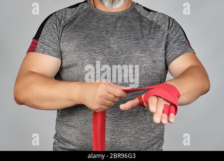 Athlet stand in grauer Kleidung und seine Hände in Rot Textil elastische Bandage wrap vor dem Training, grauer Hintergrund Stockfoto