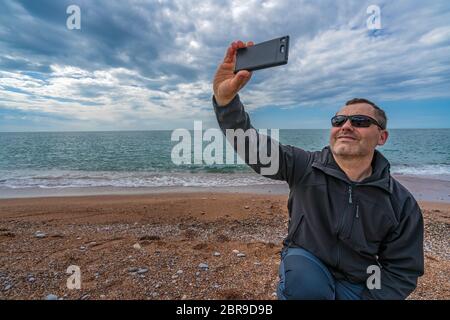Kaukasische Mann mit Sonnenbrille, selfie mit Handy am Strand im Herbst Stockfoto