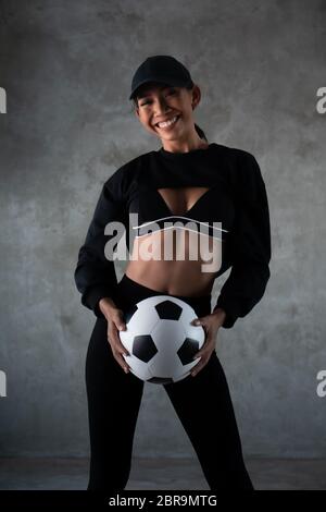 Porträt von schönen jungen lächelnden Frau in Sportkleidung hält Fußball isoliert über Beton Hintergrund Stockfoto