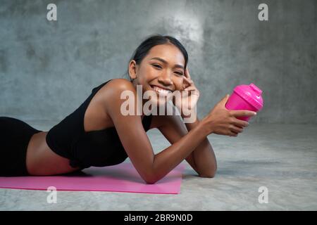 Glückliche gesunde lächelnde Frau hält Protein Shake entspannend auf der rosa Yoga-Matte im Fitnessstudio. Frau in Sportkleidung posiert im Fitnessstudio Stockfoto