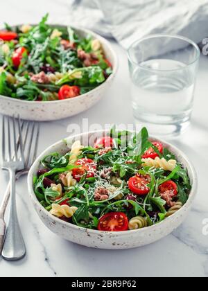 Warmer Salat mit Thunfisch, Rucola, Tomaten, roten Bohnen, Nudeln. Idee und Rezept für gesundes Mittag- oder Abendessen. Zwei Schüsseln mit warmen Salaten auf Marmor Tischplatte. Stockfoto