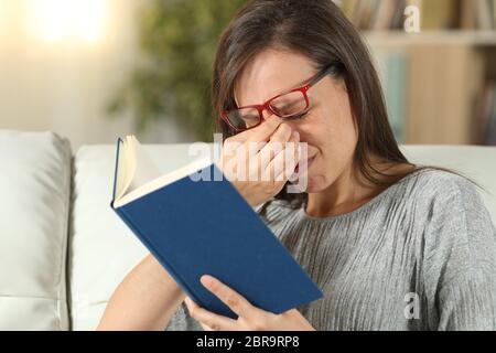 Frau mit Brille leiden Überanstrengung der Augen beim Lesen eines Buches auf einer Couch im Wohnzimmer zu Hause sitzen Stockfoto