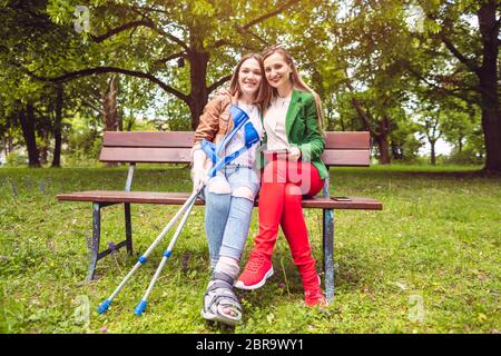 Zwei Frauen, eine gesund und eine mit verstauchten Füßen, auf einer Bank im Park Stockfoto