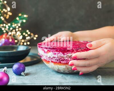 Frauenhände mit mehrschichtigem Salathering unter einem Pelzmantel auf Festtafel. Traditioneller russischer Salat mit Hering und Gemüse in Glasschüssel. Kopie sp Stockfoto