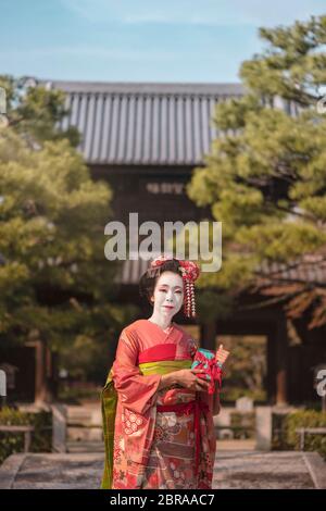 Maiko in einem Kimono posiert in Kyoto auf einem steinernen Brücke vor dem Tor von einem traditionellen Japanischen kennin Tempel umgeben von Pinien. Stockfoto