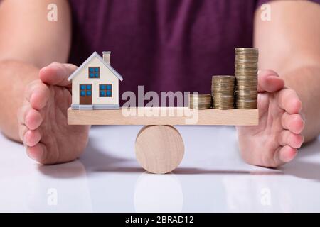 Ist eine Person, die schützende Hand Balance zwischen Haus und Geld Münzen auf einer hölzernen Wippe Stockfoto