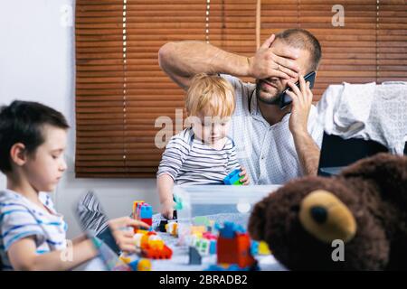 Papa mit zwei kleinen Jungen auf den Knien versucht zu Hause zu lachen und bittet die Kinder, ruhig zu sein. Ein junger Mann kümmert sich um Kinder und arbeitet an einem Komput Stockfoto