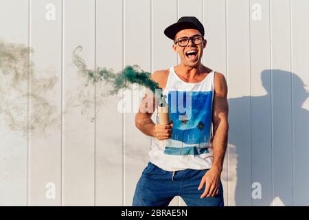 Stilvolle lächelnden Mann in Brillen Holding grünen Rauch Bombe, in der Nähe der Wand stehend, im Freien. In modischem Hemd, Hose und Mütze bekleidet. Stockfoto
