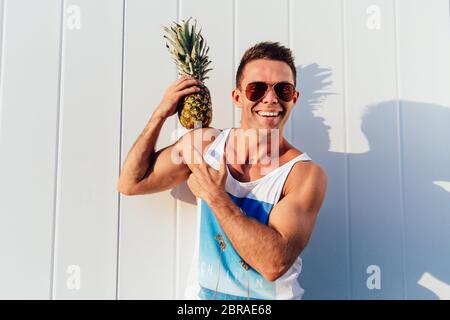 Fröhliche attraktiver Mann in Sonnenbrille hält eine Ananas auf die Schulter und zeigt auf, mit Blick auf die Kamera und lächelnd. Außerhalb. Stockfoto