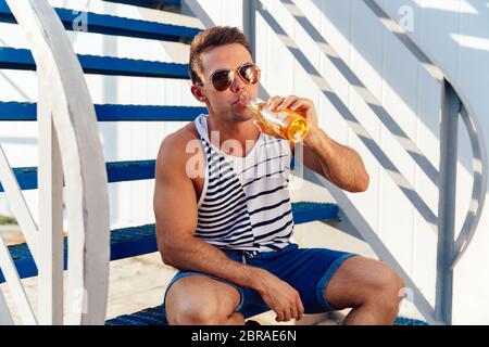 Stilvolle junger Mann in Sonnenbrille trinken ein kaltes Bier, während auf Treppen an den sonnigen Tag sitzen, im Freien. Stockfoto