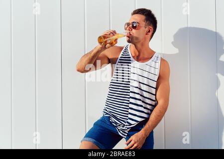 Muskulöse attraktiver Mann in Sonnenbrille trinken kaltes Bier, genießen die heißen Sommertage, im Freien. Im stilvollen Leibchen und Hosen gekleidet. Profil. Stockfoto